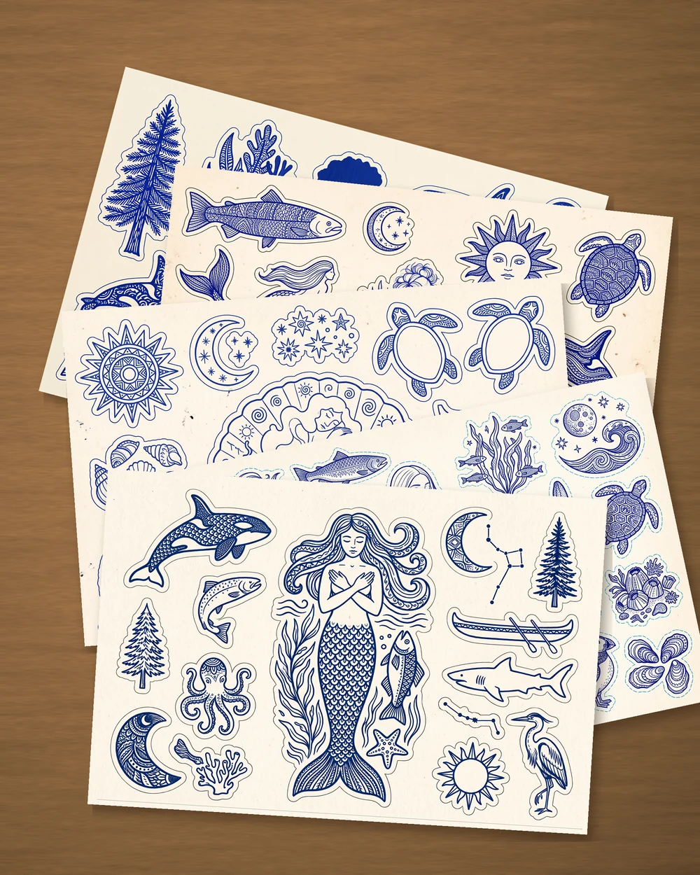 Five sticker sheets fanned across a wooden table, each printed in one blue on cream: mermaids, orcas, salmon, an octopus, sea turtles, a heron, a raven, cedar trees, kelp, coral, shells, moons, constellations and a sun with a face